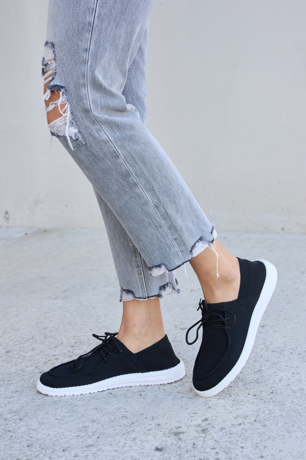 A person wearing ripped jeans and Forever Link Flat Round Toe Lace-Up Sneakers with white soles stands on a concrete surface.