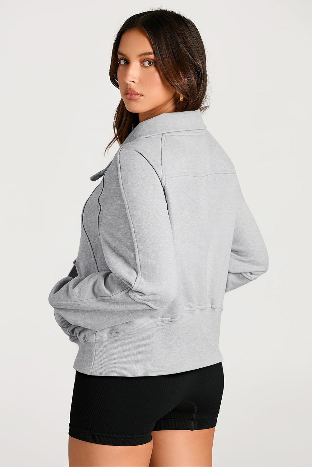 A woman with long, dark hair stands sideways, wearing a Light Grey Quarter Zip Stand Neck Kangaroo Pocket Sweatshirt that is perfect for layering, along with black shorts. She looks at the camera against a plain background, making this outfit ideal for casual occasions.