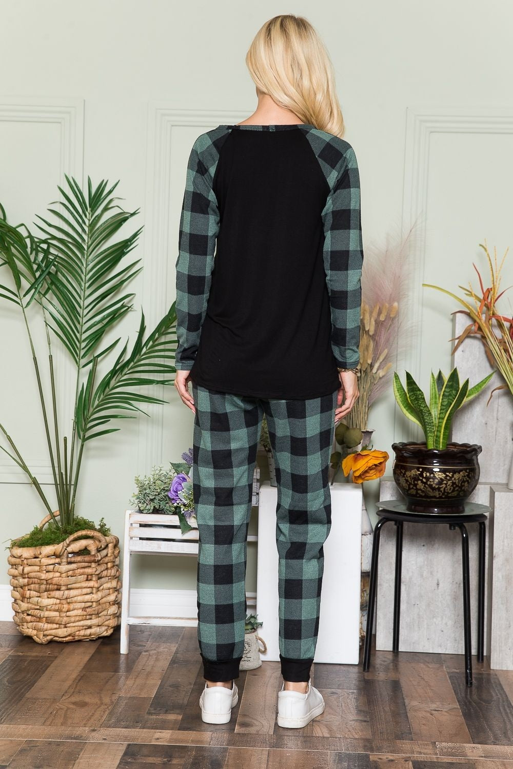 An individual wearing a Celeste Plaid Long Sleeve T-Shirt with Pockets stands indoors on a wooden floor, next to potted plants and a wicker basket.