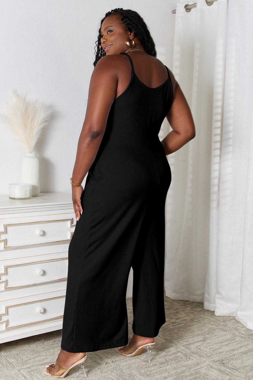 A woman in a stylish Basic Bae Full Size Spaghetti Strap V-Neck Jumpsuit stands indoors near a white dresser adorned with decorative items, smiling with one hand in her pocket.