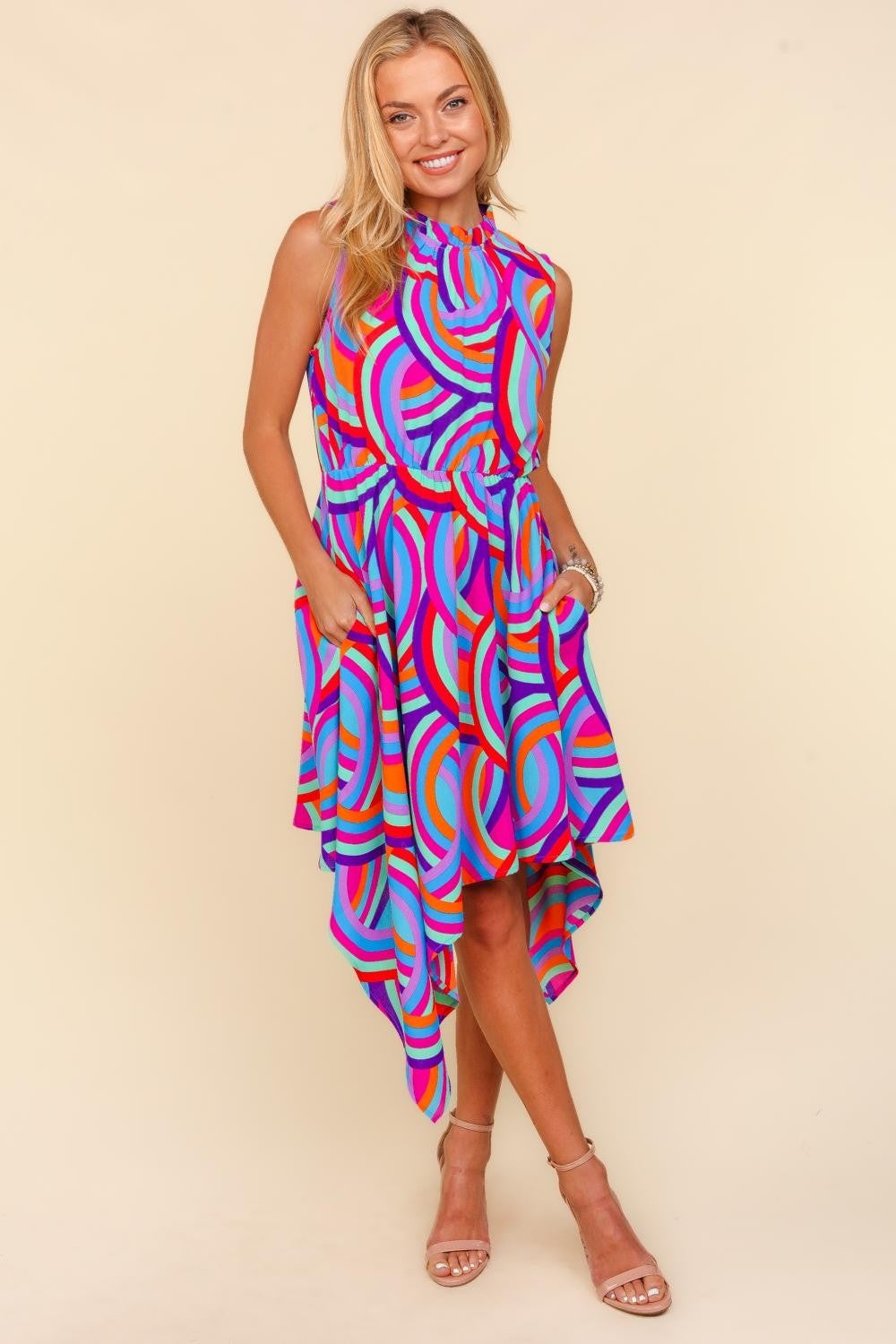 Against a beige backdrop, an individual models the Haptics Full Size Mock Neck Sleeveless Printed Dress, which features an eye-catching asymmetrical pattern. The lively blend of pink, blue, orange, and purple swirls crafts a captivating visual symphony.