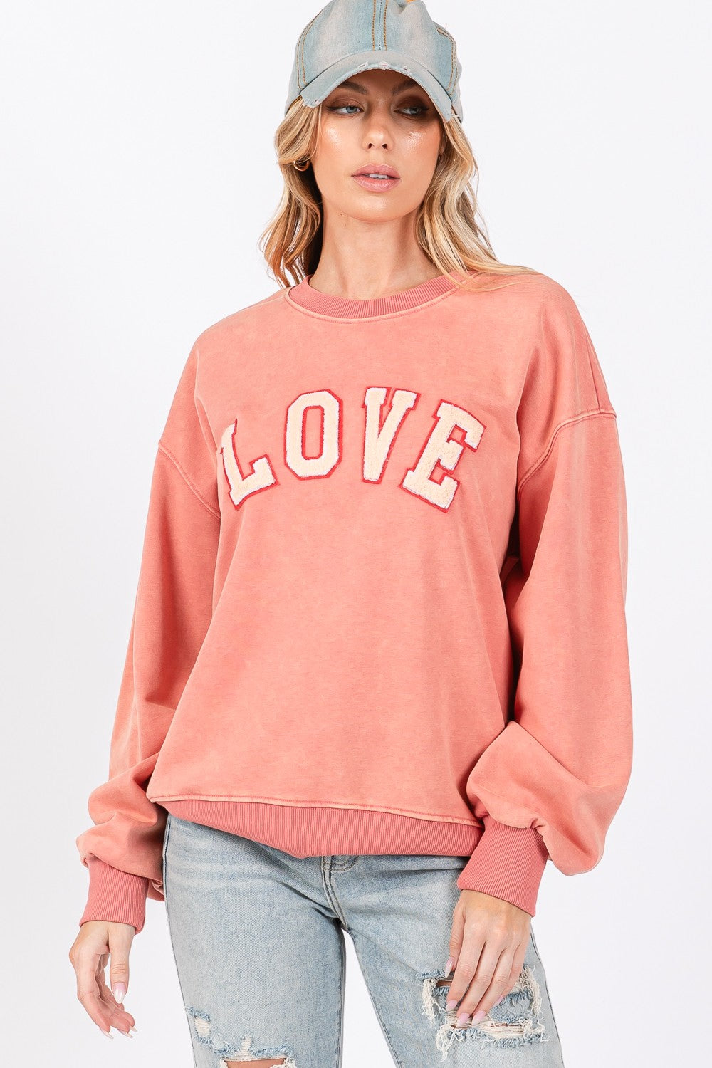 A person with long hair wears a cap, an oversized pink SAGE + FIG LOVE Path Applique Drop Shoulder Sweatshirt, and ripped jeans, standing against a plain background.