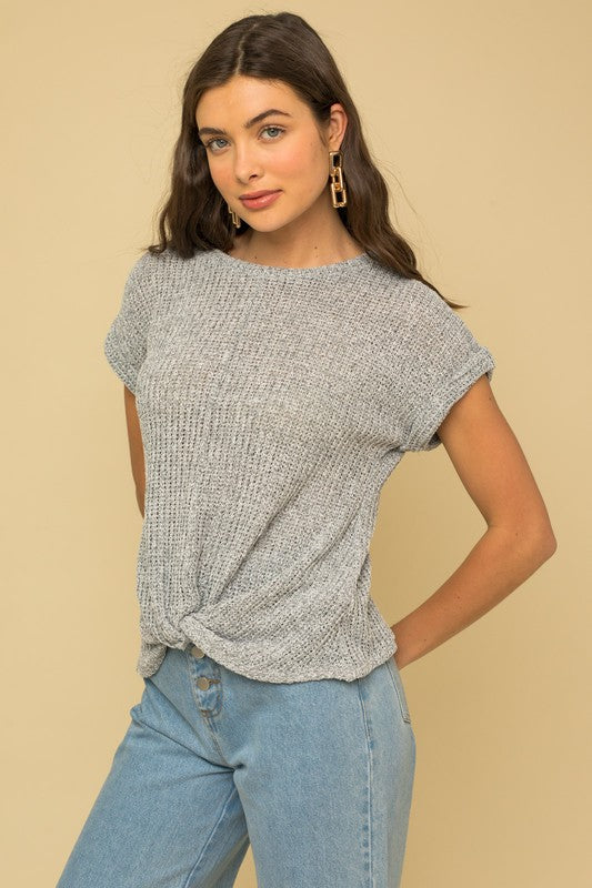 A person is seen wearing a Short Sleeve Bottom Twist Top in gray, paired with light blue jeans, standing against a beige background. They have long, wavy hair and are accessorized with large, geometric earrings.