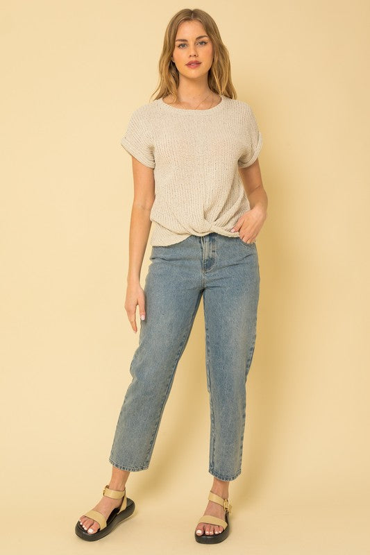 A person is seen wearing a Short Sleeve Bottom Twist Top in gray, paired with light blue jeans, standing against a beige background. They have long, wavy hair and are accessorized with large, geometric earrings.