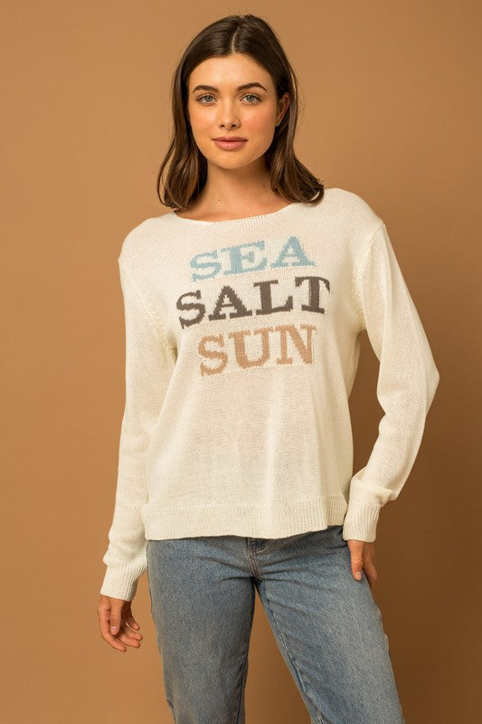 Wearing a Round Neck Long Sleeve Sea Salt Sun Sweater, beige shorts, and a straw hat, a person stands gracefully against a cream fabric backdrop, exuding coastal charm.