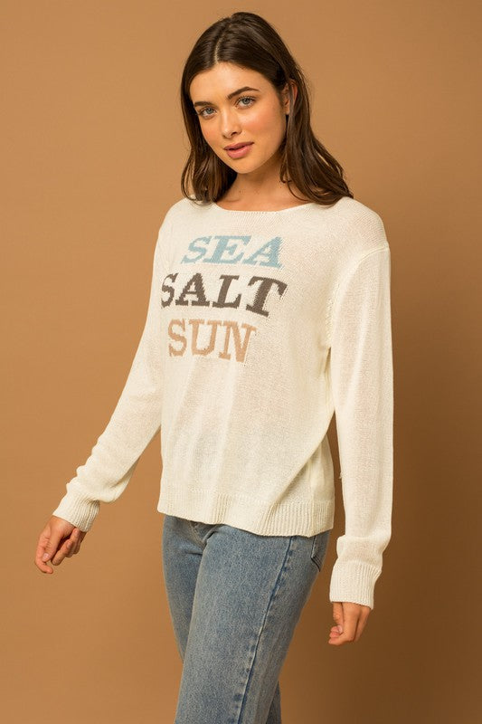 Wearing a Round Neck Long Sleeve Sea Salt Sun Sweater, beige shorts, and a straw hat, a person stands gracefully against a cream fabric backdrop, exuding coastal charm.