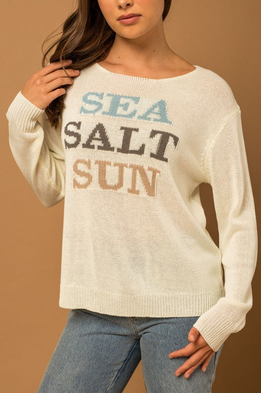 Wearing a Round Neck Long Sleeve Sea Salt Sun Sweater, beige shorts, and a straw hat, a person stands gracefully against a cream fabric backdrop, exuding coastal charm.