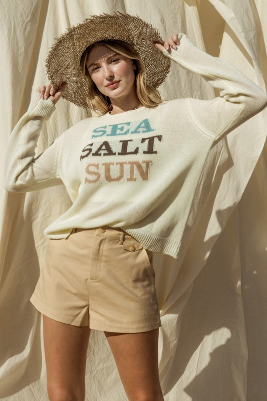 Wearing a Round Neck Long Sleeve Sea Salt Sun Sweater, beige shorts, and a straw hat, a person stands gracefully against a cream fabric backdrop, exuding coastal charm.