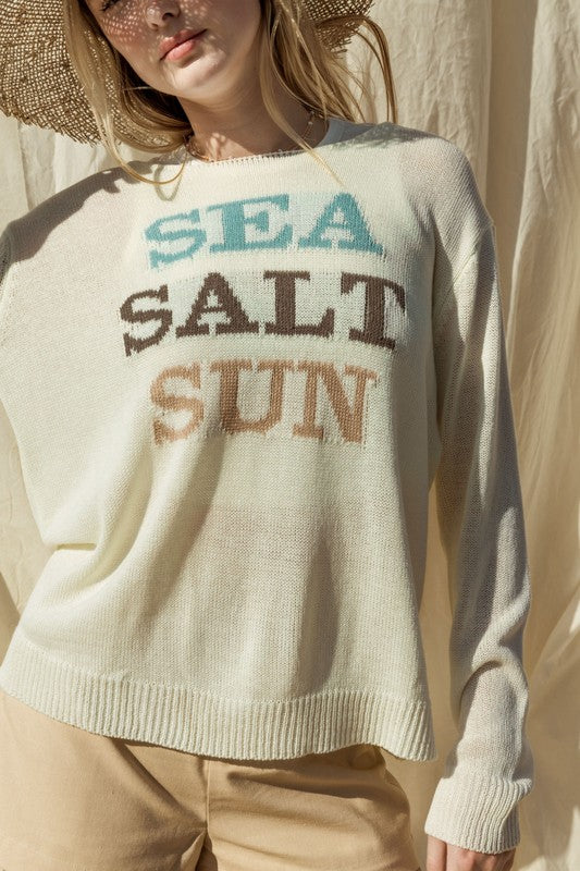 Wearing a Round Neck Long Sleeve Sea Salt Sun Sweater, beige shorts, and a straw hat, a person stands gracefully against a cream fabric backdrop, exuding coastal charm.