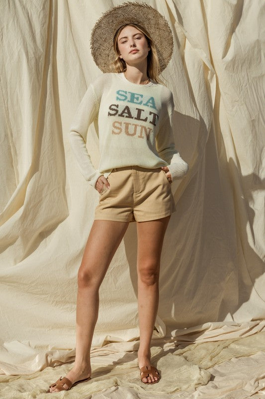 Wearing a Round Neck Long Sleeve Sea Salt Sun Sweater, beige shorts, and a straw hat, a person stands gracefully against a cream fabric backdrop, exuding coastal charm.