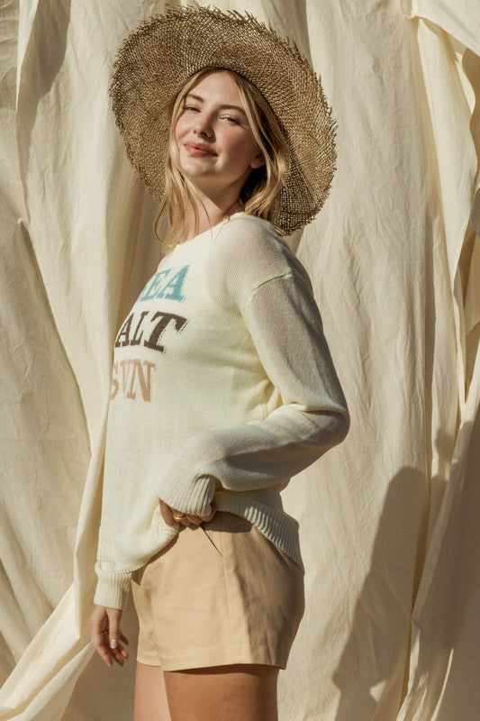 Wearing a Round Neck Long Sleeve Sea Salt Sun Sweater, beige shorts, and a straw hat, a person stands gracefully against a cream fabric backdrop, exuding coastal charm.