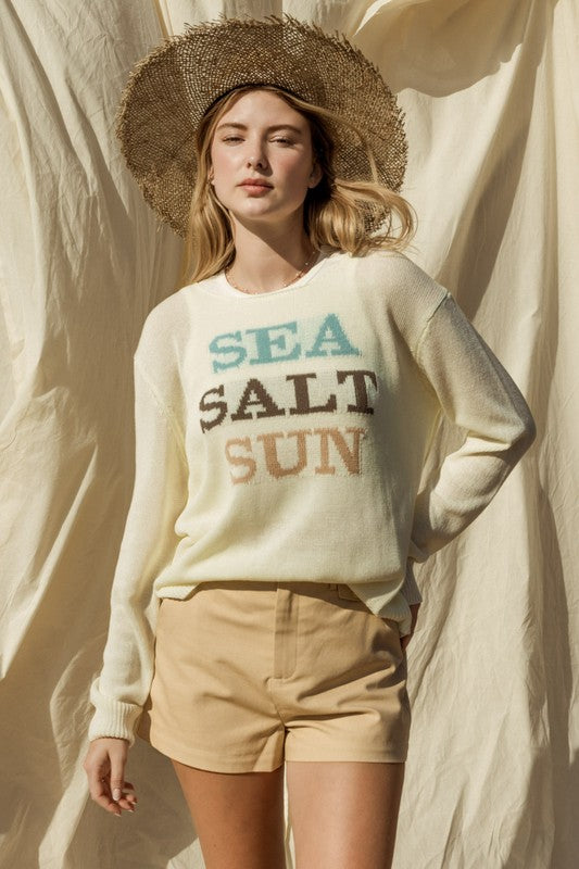 Wearing a Round Neck Long Sleeve Sea Salt Sun Sweater, beige shorts, and a straw hat, a person stands gracefully against a cream fabric backdrop, exuding coastal charm.