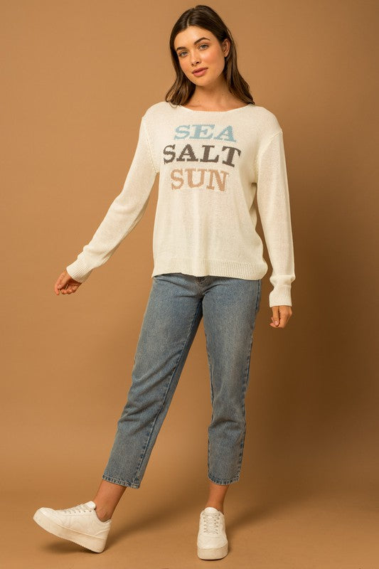 Wearing a Round Neck Long Sleeve Sea Salt Sun Sweater, beige shorts, and a straw hat, a person stands gracefully against a cream fabric backdrop, exuding coastal charm.