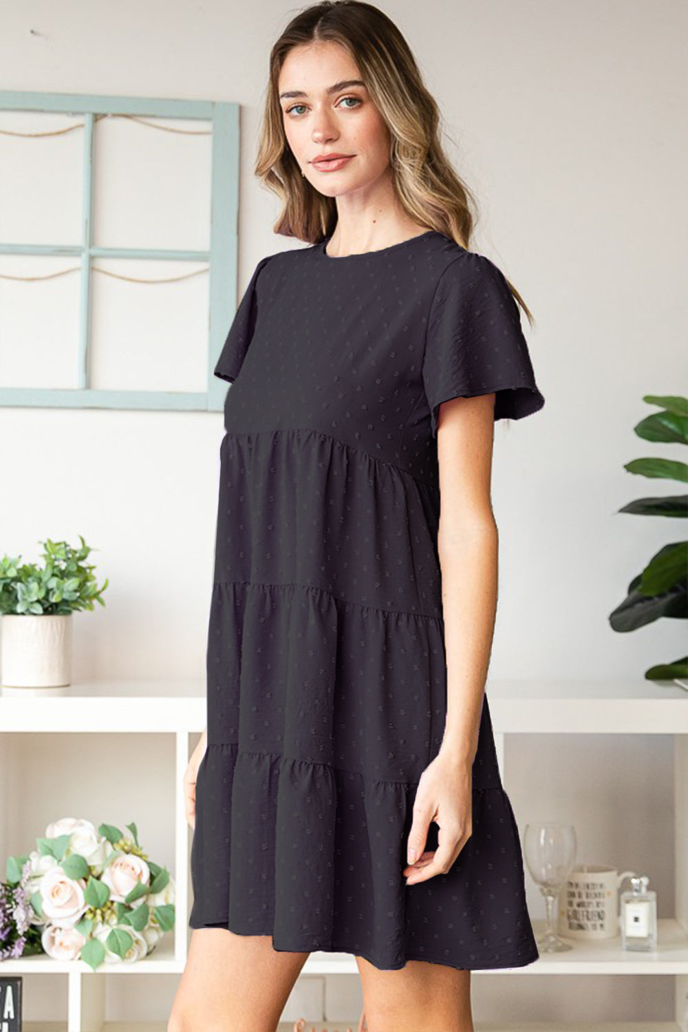 A woman elegantly poses indoors wearing the Heimish Swiss Dot Short Sleeve Tiered Dress in chic black, showcasing its stylish tiered design. The lush plants and tidy shelves create a perfect backdrop for her ensemble.