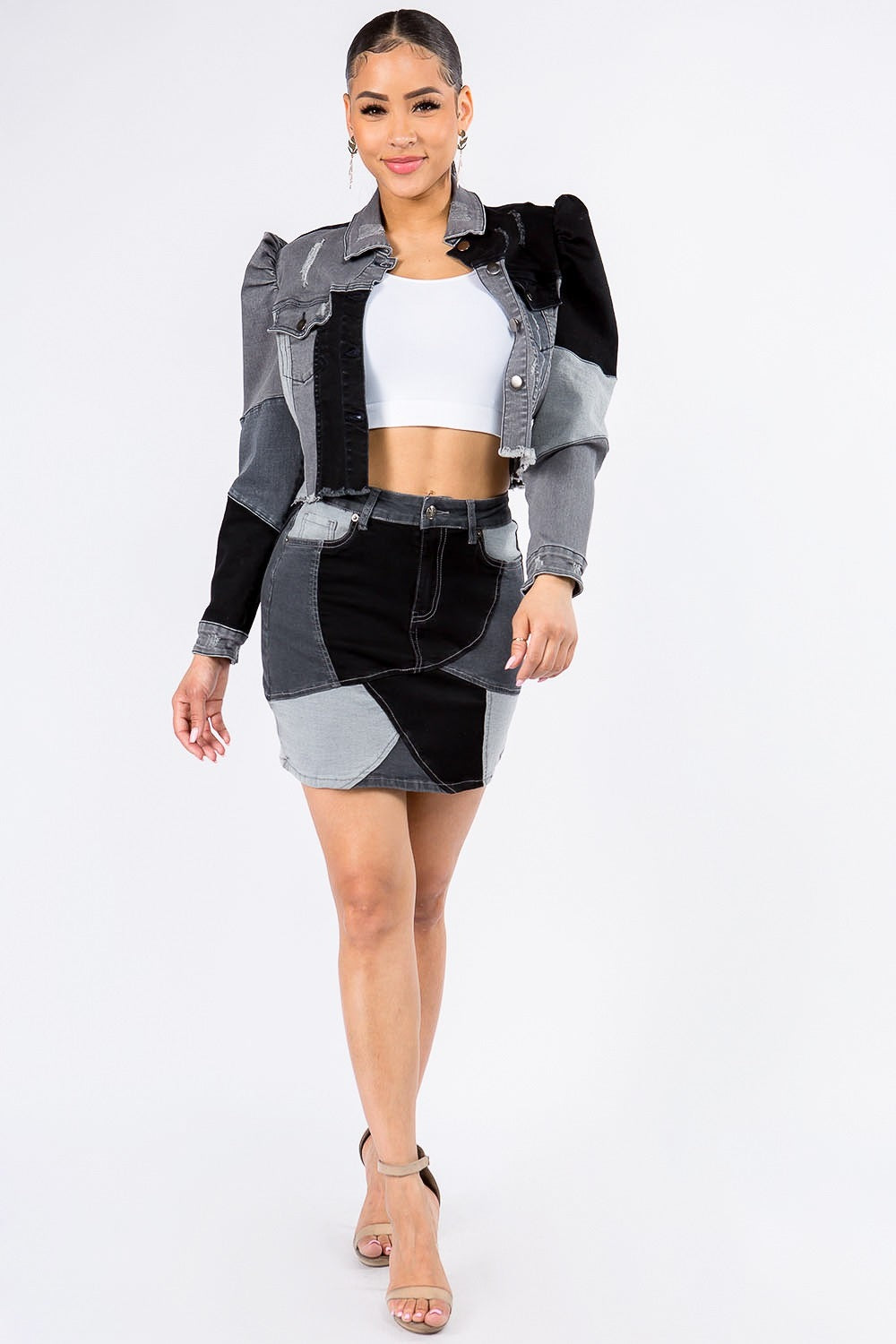 A woman is wearing the American Bazi Button Up Cropped Patchwork Denim Jacket, featuring puff shoulders and a raw hem, paired with a matching skirt in black, grey, and white. Underneath, she sports a white crop top while standing against a plain white background.