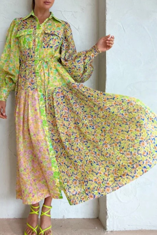 A person wearing a stunning Floral Long Sleeve Maxi Dress with a belt and matching green strappy sandals stands against a white wall, holding out part of the dress to show its flowy fabric.