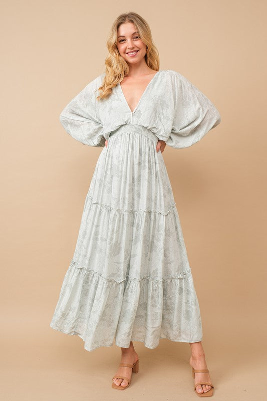 A woman in a flowy, light blue Jacquard Floral Dolman Maxi Dress with long, voluminous sleeves stands against a beige background, smiling with her hands on her hips. She wears tan, open-toed heels.