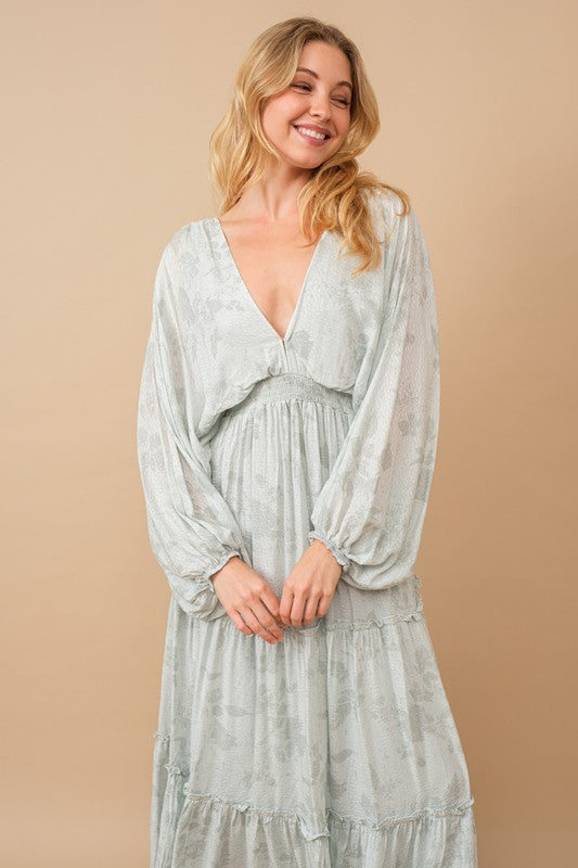 A person poses in a light green Jacquard Floral Dolman Maxi Dress with a deep V-neck and puffed sleeves. They stand against a beige background, wearing tan high-heeled sandals.