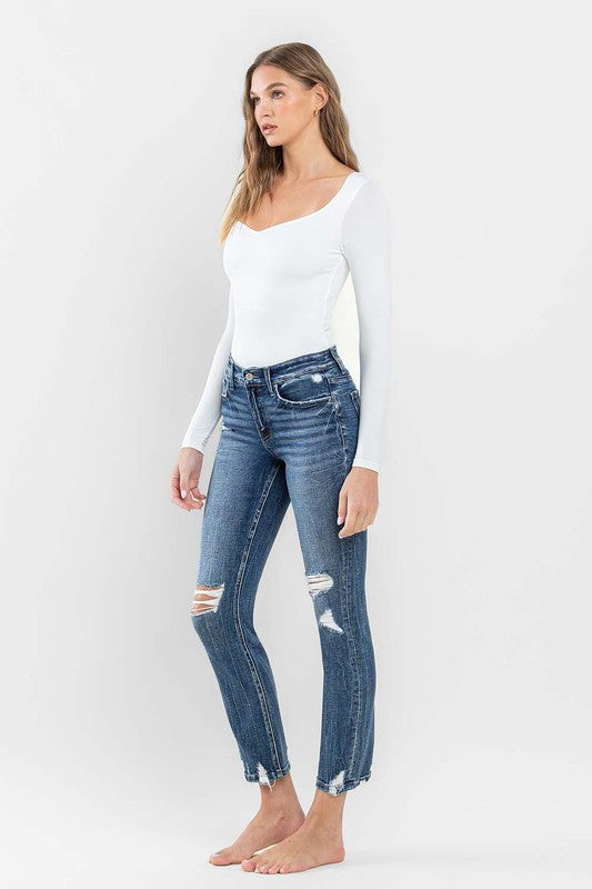 A person stands barefoot against a plain background, wearing a white long-sleeve top and the stylish Mid Rise Distressed Crop Slim Straight Jeans that perfectly capture casual elegance.