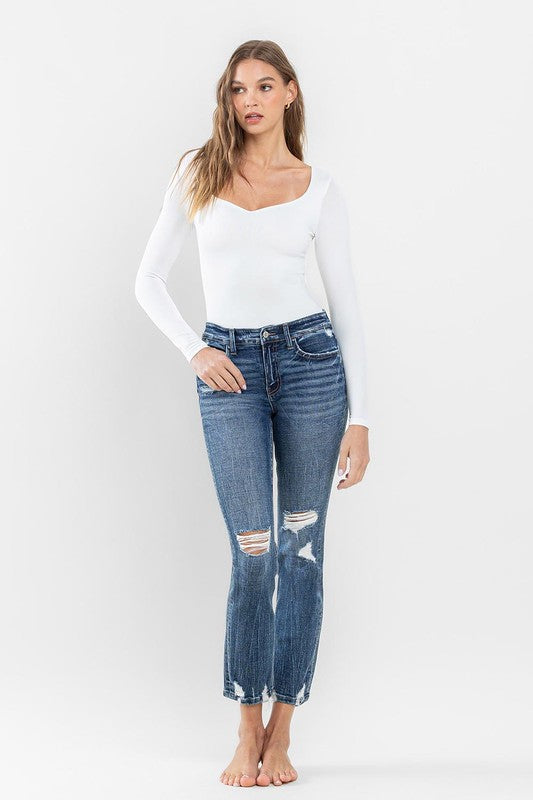 A person stands barefoot against a plain background, wearing a white long-sleeve top and the stylish Mid Rise Distressed Crop Slim Straight Jeans that perfectly capture casual elegance.