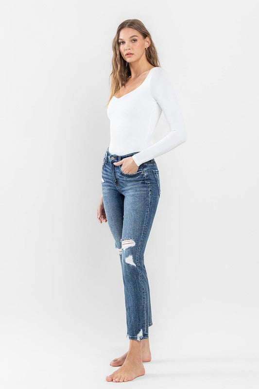 A person stands barefoot against a plain background, wearing a white long-sleeve top and the stylish Mid Rise Distressed Crop Slim Straight Jeans that perfectly capture casual elegance.