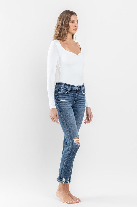 A person stands barefoot against a plain background, wearing a white long-sleeve top and the stylish Mid Rise Distressed Crop Slim Straight Jeans that perfectly capture casual elegance.