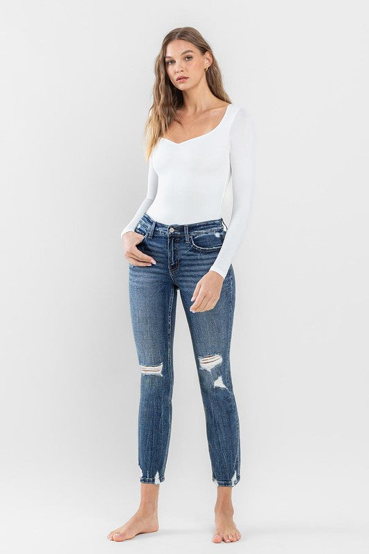 A person stands barefoot against a plain background, wearing a white long-sleeve top and the stylish Mid Rise Distressed Crop Slim Straight Jeans that perfectly capture casual elegance.