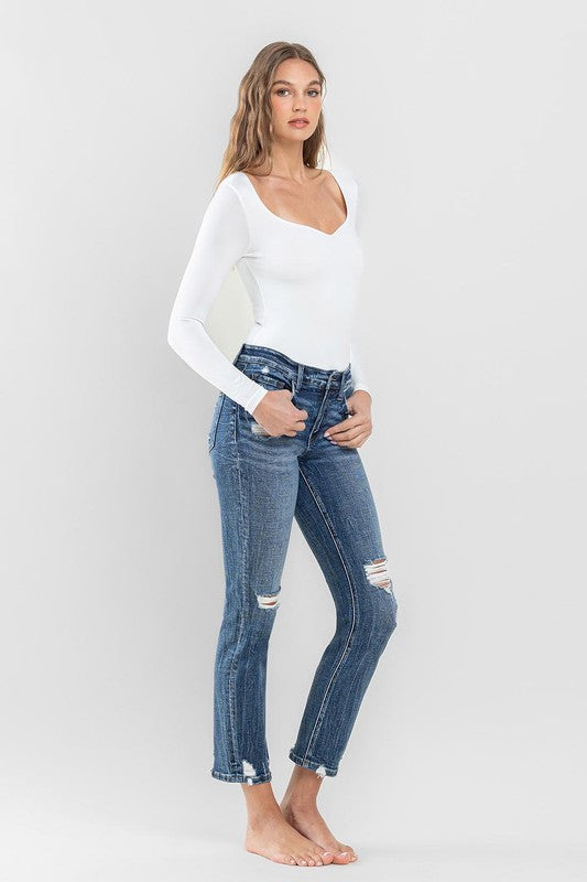 A person stands barefoot against a plain background, wearing a white long-sleeve top and the stylish Mid Rise Distressed Crop Slim Straight Jeans that perfectly capture casual elegance.