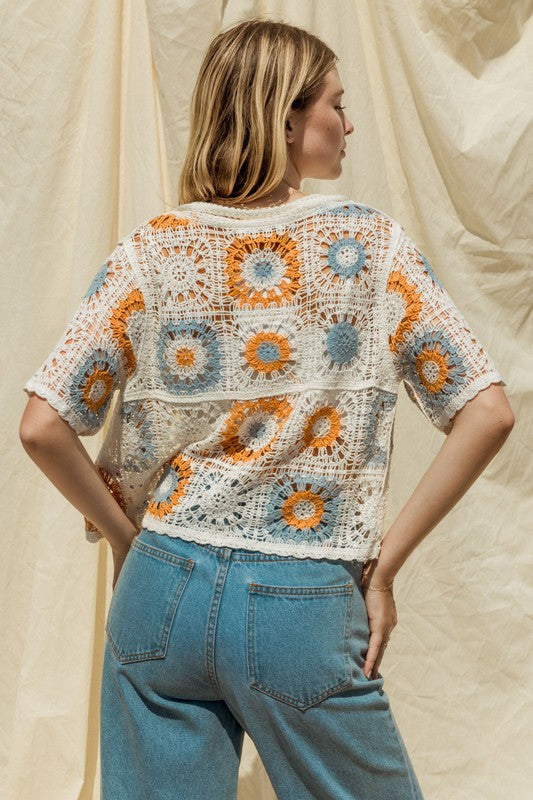 A woman adorned in a Short Sleeve Button Front Crochet Top featuring multicolored floral patterns and paired with blue jeans, exuding a Bohemian flair, stands in front of a neutral backdrop.