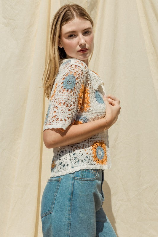 A woman adorned in a Short Sleeve Button Front Crochet Top featuring multicolored floral patterns and paired with blue jeans, exuding a Bohemian flair, stands in front of a neutral backdrop.