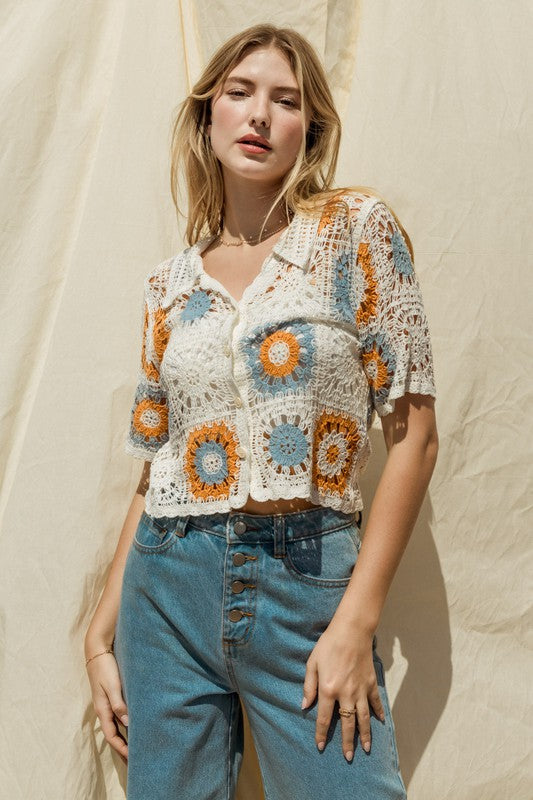 A woman adorned in a Short Sleeve Button Front Crochet Top featuring multicolored floral patterns and paired with blue jeans, exuding a Bohemian flair, stands in front of a neutral backdrop.