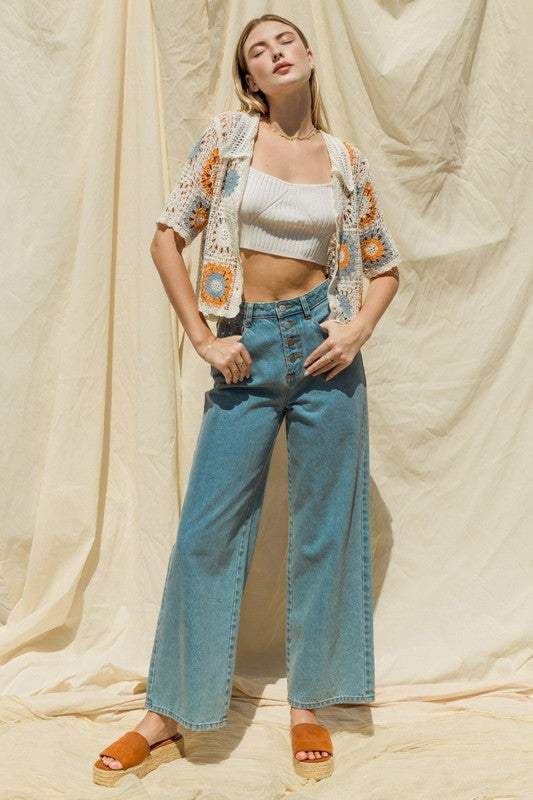 A woman adorned in a Short Sleeve Button Front Crochet Top featuring multicolored floral patterns and paired with blue jeans, exuding a Bohemian flair, stands in front of a neutral backdrop.