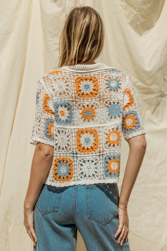 A woman adorned in a Short Sleeve Button Front Crochet Top featuring multicolored floral patterns and paired with blue jeans, exuding a Bohemian flair, stands in front of a neutral backdrop.