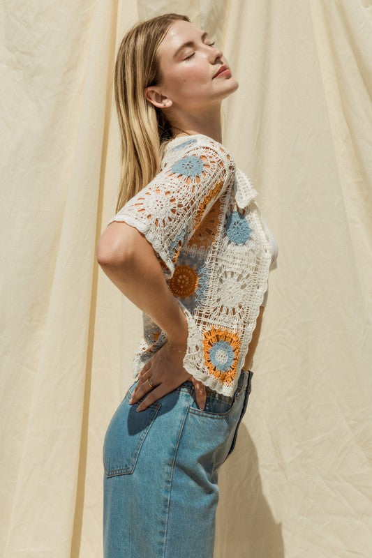 A woman adorned in a Short Sleeve Button Front Crochet Top featuring multicolored floral patterns and paired with blue jeans, exuding a Bohemian flair, stands in front of a neutral backdrop.