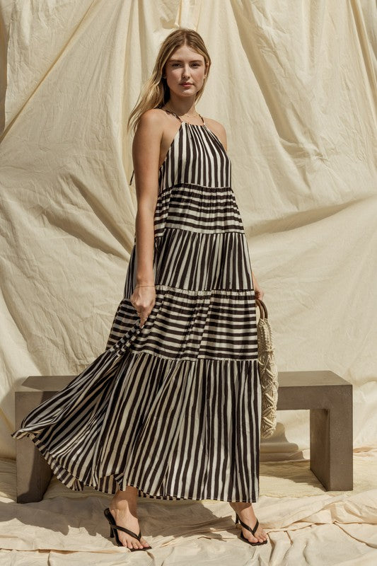 A woman in a Sleeveless Stripe Maxi Tiered Dress and hat stands by a suitcase outside, with mountains and palm trees in the background.