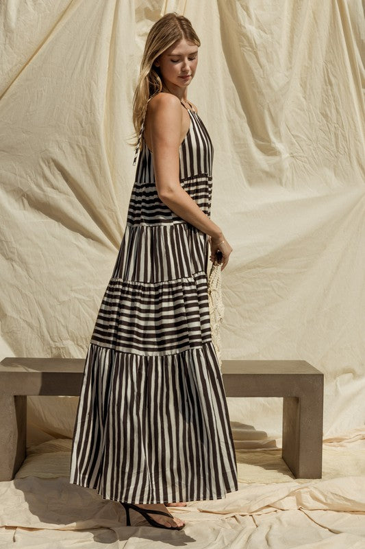 A woman in a Sleeveless Stripe Maxi Tiered Dress and hat stands by a suitcase outside, with mountains and palm trees in the background.