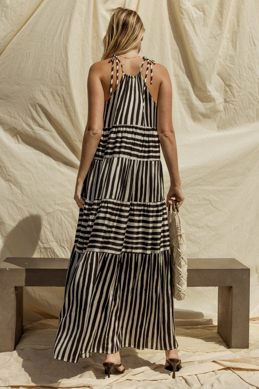 A woman in a Sleeveless Stripe Maxi Tiered Dress and hat stands by a suitcase outside, with mountains and palm trees in the background.