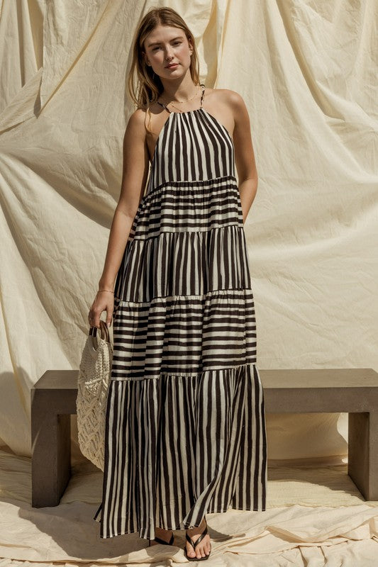 A woman in a Sleeveless Stripe Maxi Tiered Dress and hat stands by a suitcase outside, with mountains and palm trees in the background.