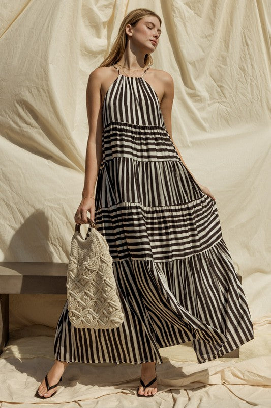 A woman in a Sleeveless Stripe Maxi Tiered Dress and hat stands by a suitcase outside, with mountains and palm trees in the background.