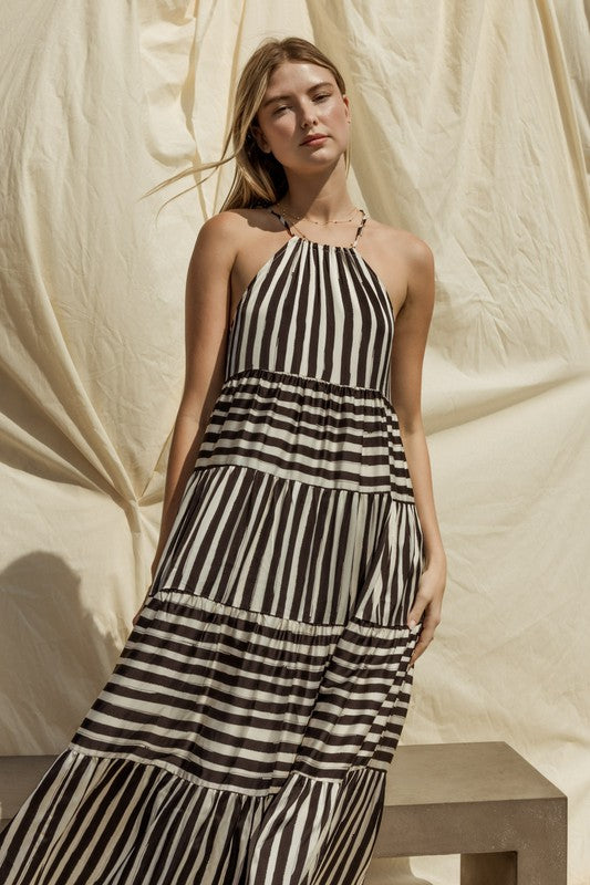 A woman in a Sleeveless Stripe Maxi Tiered Dress and hat stands by a suitcase outside, with mountains and palm trees in the background.