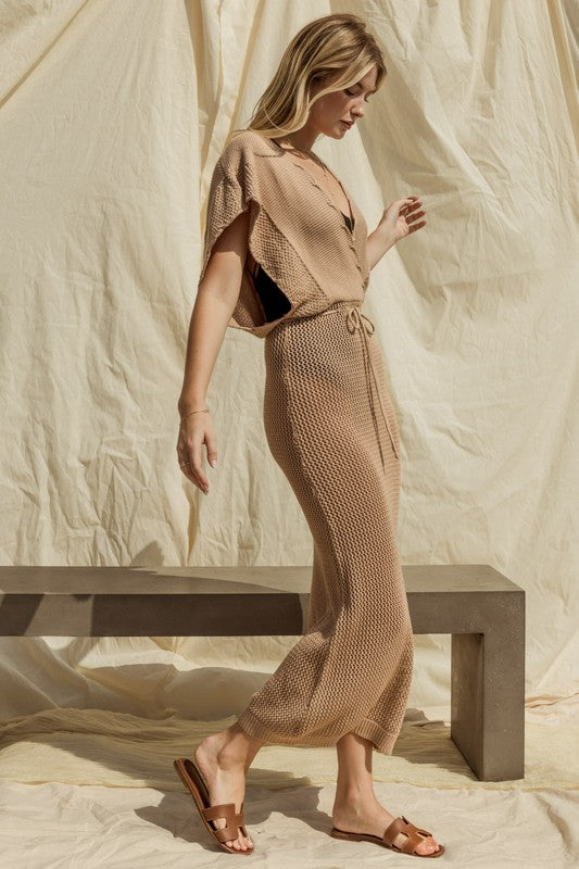 Wearing the chic Open Sweater Cover-Up Dress, a woman sits elegantly on a concrete bench against a matching beige fabric backdrop, stylishly gazing to her left.