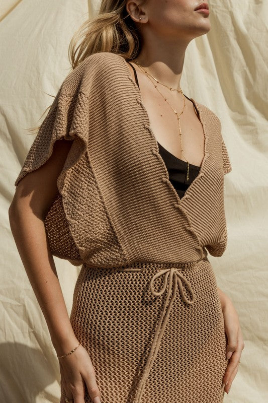 Wearing the chic Open Sweater Cover-Up Dress, a woman sits elegantly on a concrete bench against a matching beige fabric backdrop, stylishly gazing to her left.