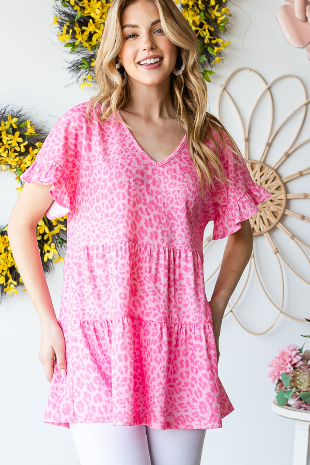 A person wearing a Heimish Full Size Leopard Ruffle Trim Short Sleeve Top stands in front of a floral wreath display.