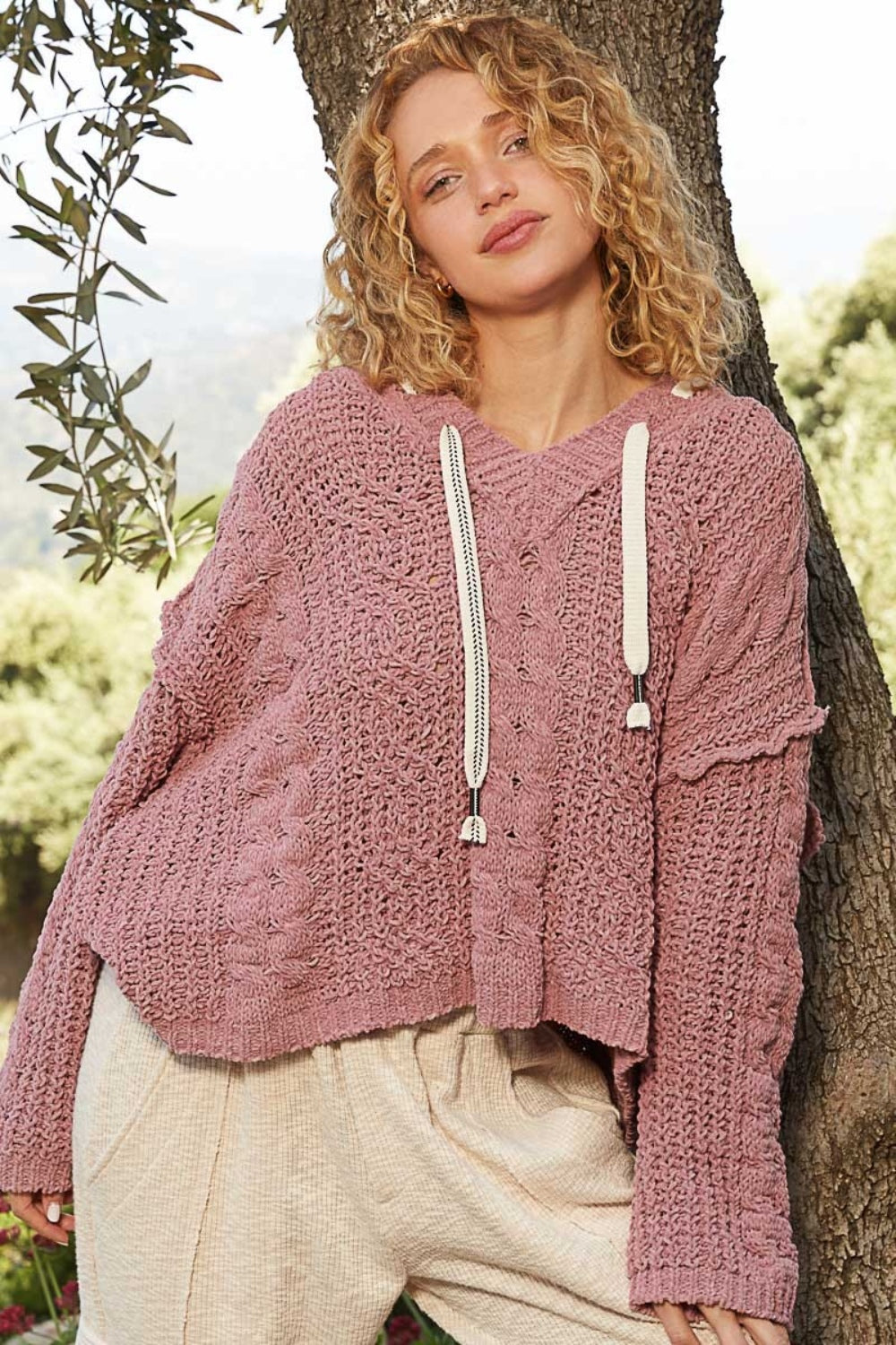 A person with curly blond hair is standing outdoors, leaning against a tree, wearing a cozy POL Cable Knit Hooded Chenille Sweater in pink and light-colored pants.