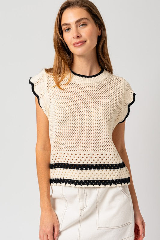 A person is wearing the Round Neck Ruffle Sleeveless Color Contrast Top, which is cream-colored with black trim and features a round neck design, paired with a white skirt, standing with hands on hips against a plain backdrop.