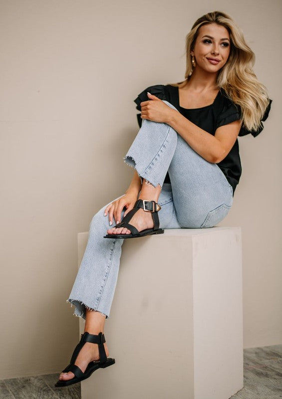 A person seated on a block wearing white pants, a patterned top, and tan AVA 2 sandals with supple leather ankle straps and a signature buckle. The person's face is not visible.