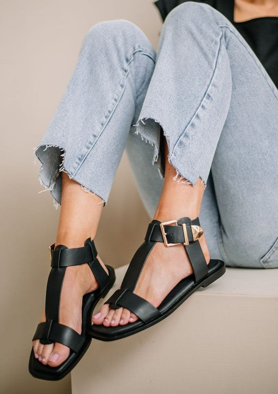 A person seated on a block wearing white pants, a patterned top, and tan AVA 2 sandals with supple leather ankle straps and a signature buckle. The person's face is not visible.