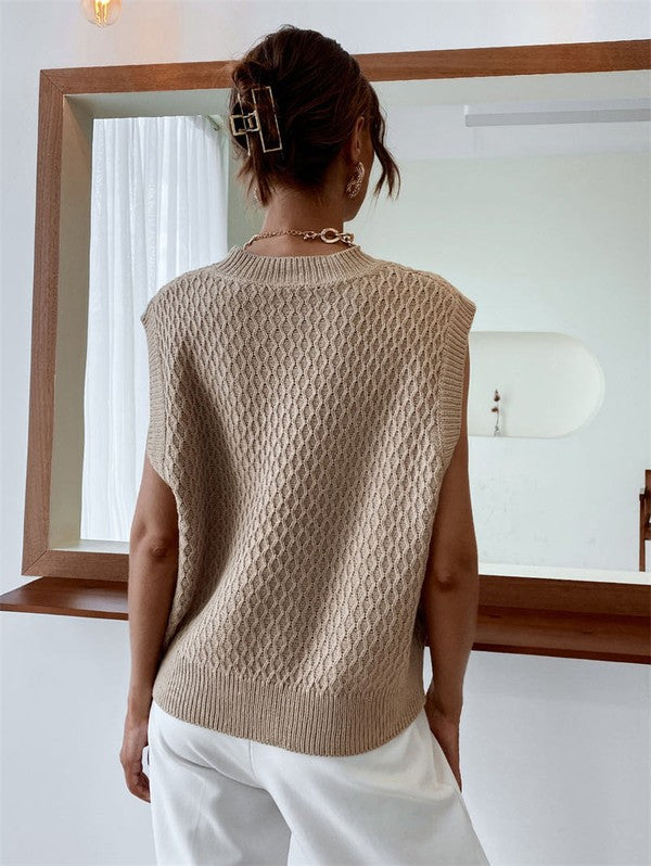 A person wearing the Knit Sweater Vest in a textured beige knit paired with white pants stands indoors, accessorized with a gold chain necklace.