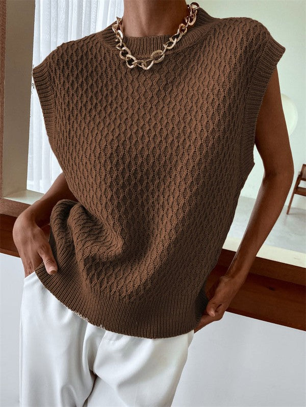 A person wearing the Knit Sweater Vest in a textured beige knit paired with white pants stands indoors, accessorized with a gold chain necklace.