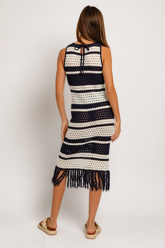 A woman in the Sleeveless Crochet Effect Sweater Dress, featuring black and white stripes with a fringe hem, holds a textured beige handbag. She pairs the outfit with beige sandals, standing against a plain white background and exuding an effortlessly chic vibe.